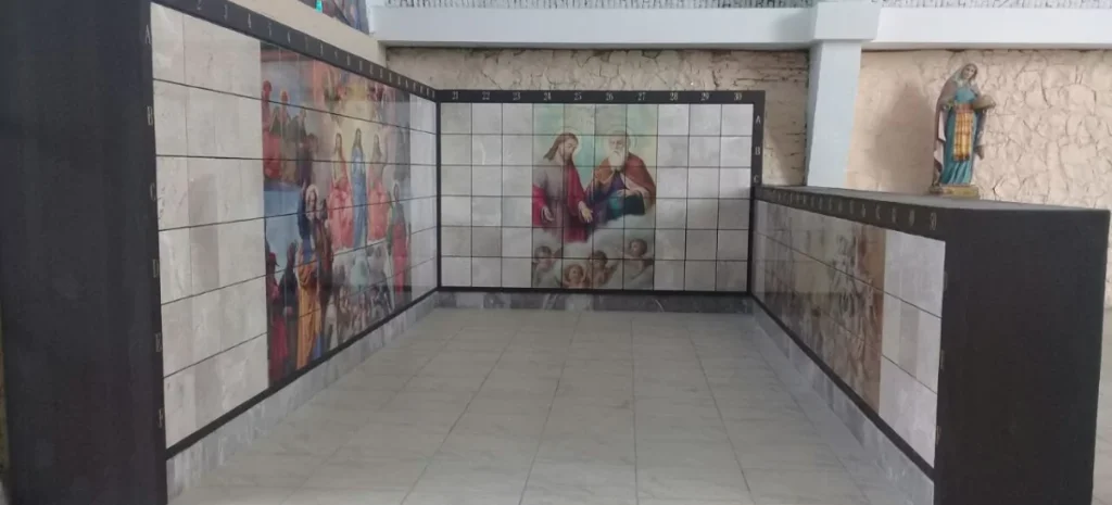 Pasillo con murales de mosaico que representan escenas religiosas, incluyendo figuras de santos y ángeles, con numeración en los bordes y una estatua de la Virgen en el extremo