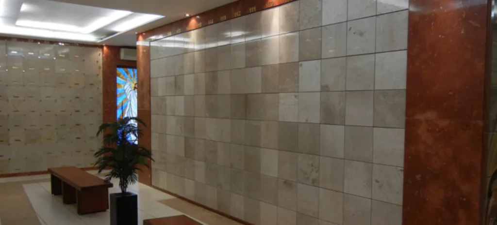 Muro de nichos en mármol beige, enmarcado con mármol rojo y luces superiores para destacar el espacio. Al fondo, una vidriera colorida aporta luminosidad al ambiente