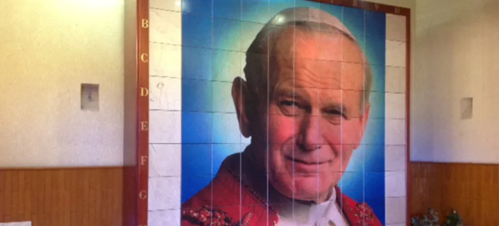Muro de nichos con retrato de San Juan Pablo II sobre fondo azul, enmarcado en mármol blanco y detalles en rojo, brindando un ambiente de respeto y devoción
